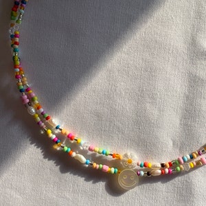 Könnte beinhalten: Mehrfarbige Perlenkette mit einem weißen Smiley-Anhänger. Die Kette besteht aus kleinen, bunten Perlen, darunter weiße Perlen, und ist für einen trendigen Look geschichtet.