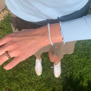 May include: A white pearl bracelet with a simple design. The bracelet is worn on a person's wrist. The person is wearing a light blue shirt and a plaid skirt.