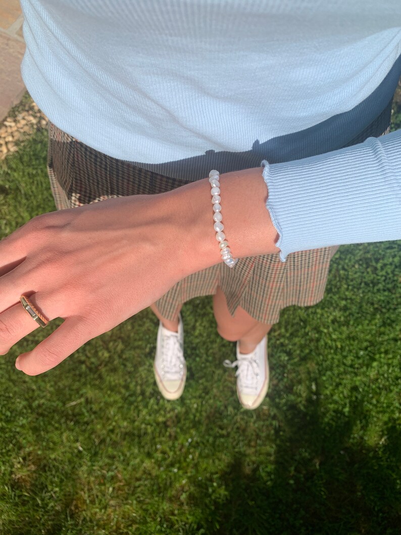 May include: A white pearl bracelet with a simple design. The bracelet is worn on a person's wrist. The person is wearing a light blue shirt and a plaid skirt.