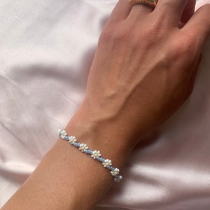 Könnte beinhalten: Ein zartes Armband mit weißen Perlen in Form von Gänseblümchen und hellblauen Perlen. Das Armband befindet sich am Handgelenk.