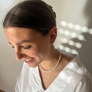 May include: A woman wearing a white button-down shirt with a pearl necklace. Her hair is pulled back with a dark hair clip.