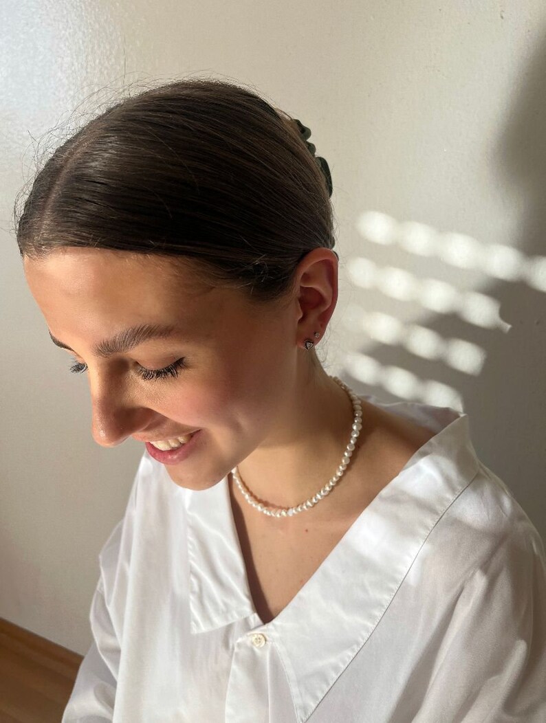 May include: A woman wearing a white button-down shirt with a pearl necklace. Her hair is pulled back with a dark hair clip.
