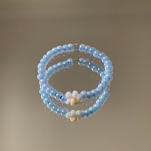 Könnte beinhalten: Ein hellblaues Perlenarmband mit zwei weißen und gelben Blumenanhängern.