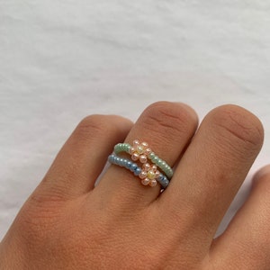 Könnte beinhalten: Zwei Perlenringe mit Blumenmustern. Ein Ring ist hellblau, der andere hellgrün. Jeder Ring hat eine Blume aus kleinen, hellfarbigen Perlen. Die Ringe befinden sich an einer Hand.