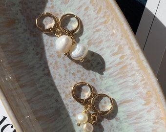 Puede incluir: Un par de pendientes de aro dorados con colgantes de perlas. Los pendientes están sobre una superficie moteada de blanco y dorado.