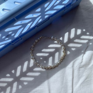 May include: A white pearl necklace with a silver clasp, lying on a white fabric surface with a blue plastic crate in the background.