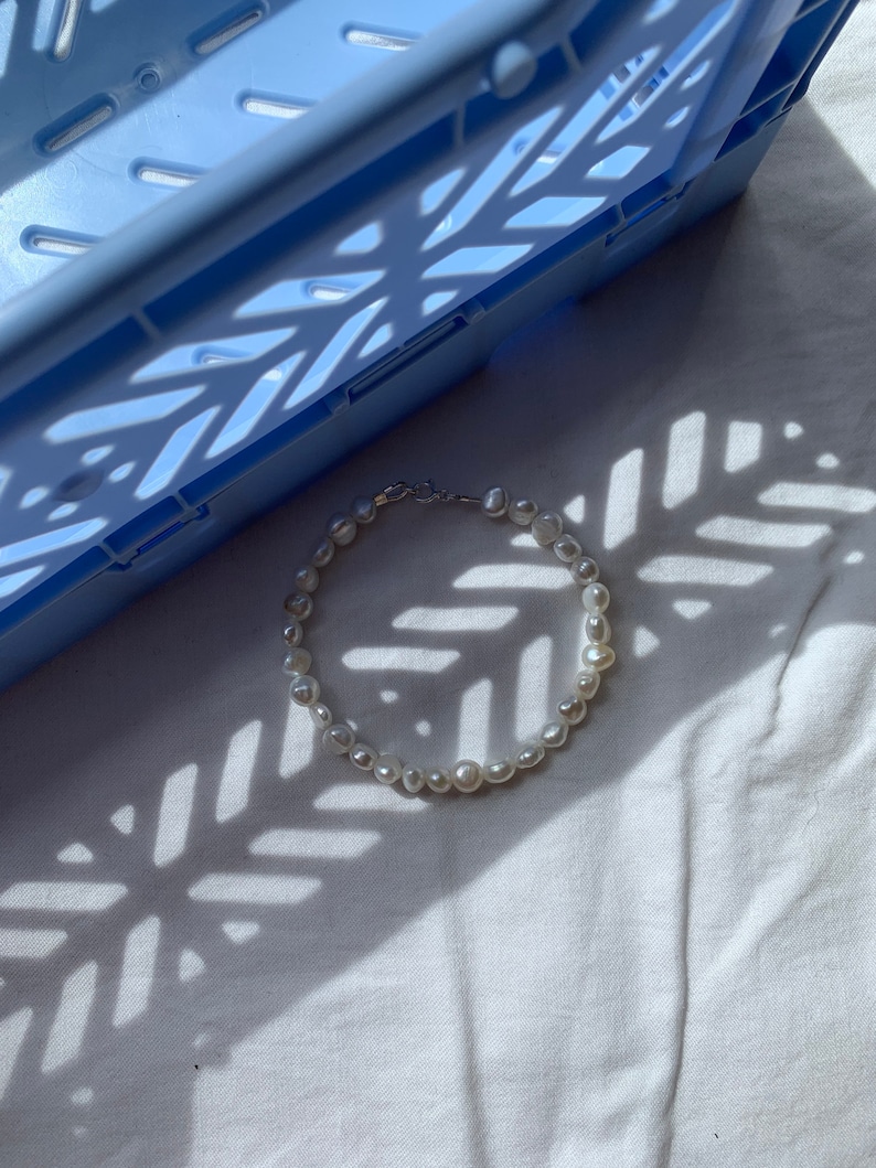 May include: A white pearl necklace with a silver clasp, lying on a white fabric surface with a blue plastic crate in the background.