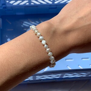 May include: A white pearl bracelet with a simple design. The bracelet is made of small, round pearls that are strung together on a thin string. The bracelet is shown on a person's wrist.