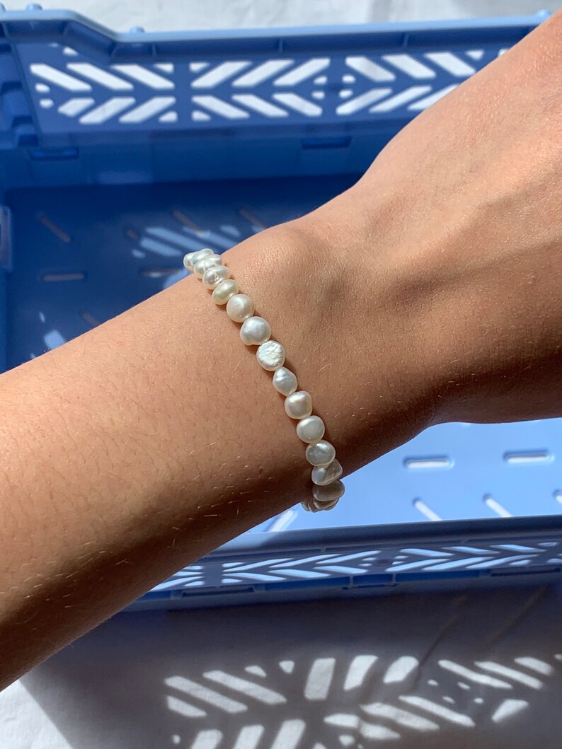May include: A white pearl bracelet with a simple design. The bracelet is made of small, round pearls that are strung together on a thin string. The bracelet is shown on a person's wrist.