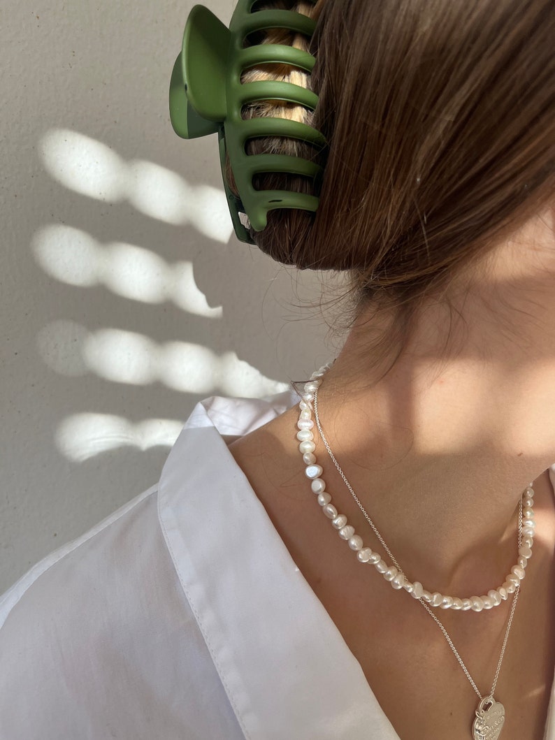 May include: A white pearl necklace with a silver heart pendant charm. The necklace is layered over a silver chain.