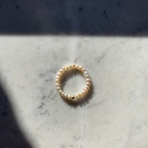 Könnte beinhalten: Ein weißes Perlenarmband mit einem goldenen Perlenakzent. Das Armband liegt auf einer weißen Marmorfläche.