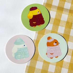 Peut inclure: Trois autocollants ronds représentant des animaux de dessins animés en vêtements d'hiver. Un chien jaune portant un manteau et un béret bordeaux sur fond vert. Un lapin blanc en manteau et chapeau bleus sur fond rose. Un ours orange en manteau et chapeau roses sur fond vert clair.