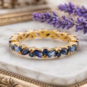 May include: A gold ring with a band of marquise-cut blue gemstones. The ring is set on a white marble surface, with a sprig of purple lavender in the background. The ring is a piece of jewelry.