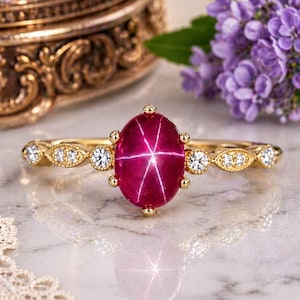 May include: A gold ring featuring a large, oval, star ruby gemstone. The ruby is a deep red color with a six-ray star pattern. The band is adorned with small, clear gemstones and decorative gold accents.