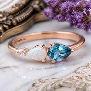 Könnte beinhalten: Ring aus Roségold mit einem birnenförmigen blauen Edelstein und einem birnenförmigen weißen Edelstein. Neben dem weißen Edelstein ist ein kleiner runder Diamant gesetzt. Der Ring liegt auf einer weißen Marmoroberfläche.