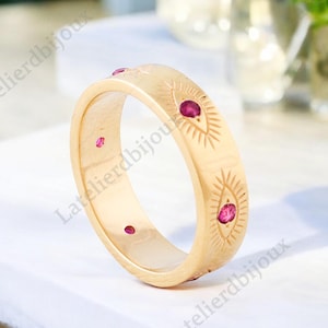 May include: Gold band ring with a repeating pattern of stylised eyes with red gemstones.