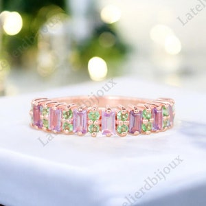 May include: A rose gold band ring with a row of rectangular pink and green gemstones. The ring is on a white surface.