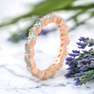 以下が含まれることがあります： ローズゴールドの永遠のバンドに、マルキーズ型のブルーのトパーズと小さなラウンドダイヤモンドが並んでいます。