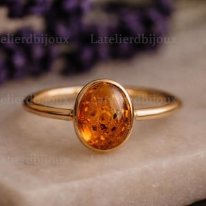 Può includere: Un anello d'oro con una pietra d'ambra ovale. L'ambra è di un colore arancione caldo e traslucido con inclusioni visibili. La fascia dell'anello è sottile e lucida, riflettendo la luce. Lo sfondo presenta fiori viola sfocati.
