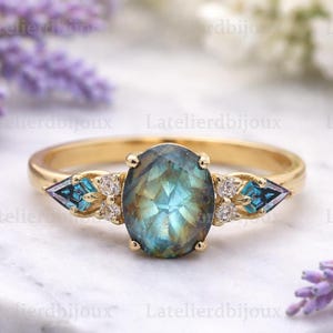 May include: A gold ring featuring an oval, iridescent gemstone as the centerpiece. The ring is flanked by blue and white accent stones. The ring is set on a white marble surface with lavender flowers in the background.