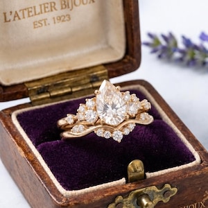 Peut inclure: Une bague en or avec une pierre centrale en forme de poire entourée de petites pierres rondes, placée dans une boîte en bois doublée de velours violet. Le couvercle porte l'inscription L'ATELIER BIJOUX EST. 1923 et l'avant indique L'ATELIER BIJOUX.