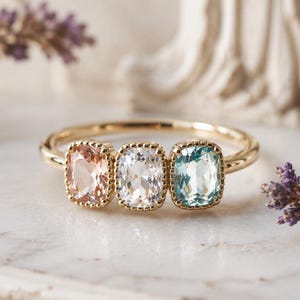 May include: A gold ring featuring three cushion-cut gemstones in peach, white, and teal. The ring is set on a white marble surface, with sprigs of purple flowers in the background. The ring is a piece of jewelry.