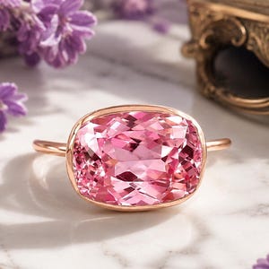 May include: A rose gold ring featuring a large, cushion-cut pink gemstone. The ring is set against a marble surface, with lavender flowers and a decorative gold-toned jewelry box in the background. The gemstone has a faceted design.
