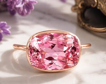 May include: A rose gold ring featuring a large, cushion-cut pink gemstone. The ring is set against a marble surface, with lavender flowers and a decorative gold-toned jewelry box in the background. The gemstone has a faceted design.