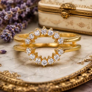 Op de afbeelding: Een goudkleurige ring met een halo van kleine, heldere edelstenen. De ring heeft een dubbelbandontwerp, waarbij de edelstenen in een cirkelvormig patroon zijn geplaatst. De ring wordt gepresenteerd op een marmeren oppervlak.