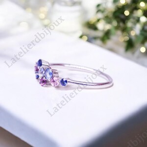 Puede incluir: Un delicado anillo de plata con un grupo de peque&ntilde;as piedras preciosas de colores. El anillo presenta un dise&ntilde;o con piedras azules, moradas y rosas, creando un aspecto brillante y elegante. El anillo est&aacute; sobre una superficie blanca.