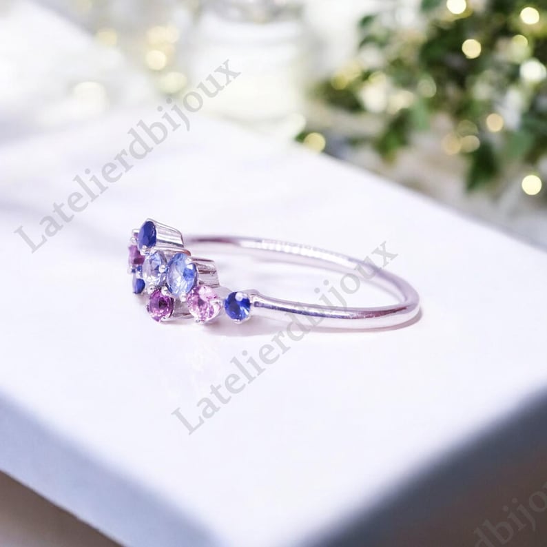 Puede incluir: Un delicado anillo de plata con un grupo de peque&ntilde;as piedras preciosas de colores. El anillo presenta un dise&ntilde;o con piedras azules, moradas y rosas, creando un aspecto brillante y elegante. El anillo est&aacute; sobre una superficie blanca.