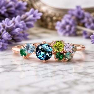 Può includere: Un anello in oro rosa con un gruppo di pietre preziose colorate. L'anello presenta una grande gemma rotonda blu scuro al centro, circondata da gemme più piccole nelle tonalità del verde, blu e viola. L'anello è posto su una superficie di marmo.