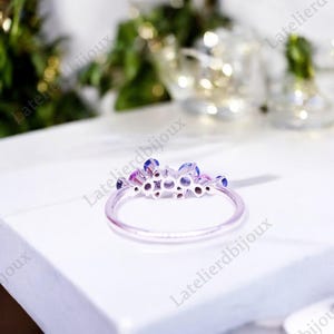 Puede incluir: Un anillo plateado con un grupo de piedras preciosas azules y rosas. El anillo tiene una banda delgada y delicada, y las piedras est&aacute;n dispuestas en un dise&ntilde;o floral o en racimo. El anillo est&aacute; sobre una superficie blanca.