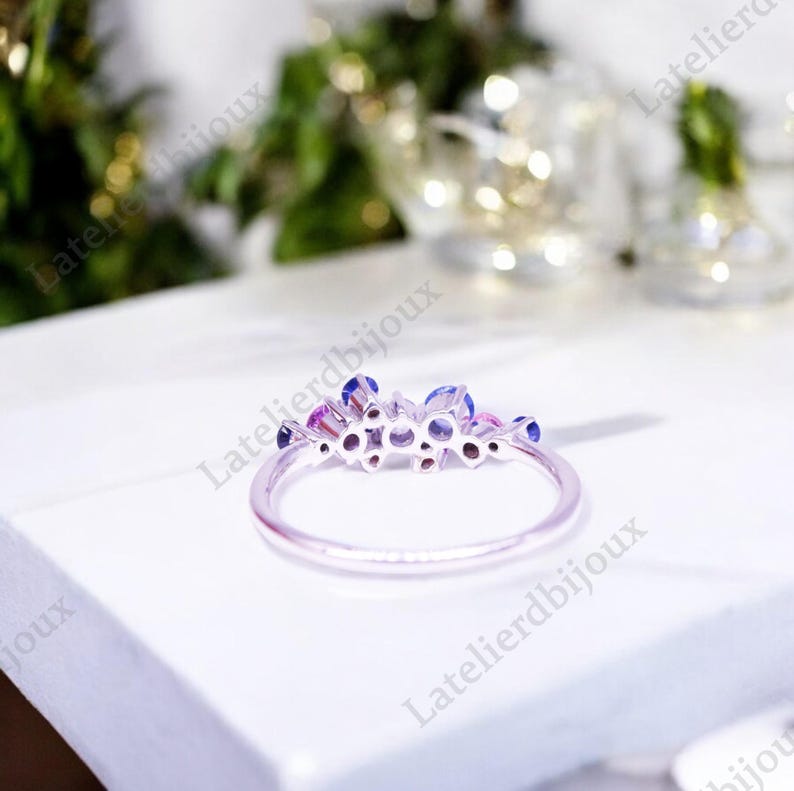 Puede incluir: Un anillo plateado con un grupo de piedras preciosas azules y rosas. El anillo tiene una banda delgada y delicada, y las piedras est&aacute;n dispuestas en un dise&ntilde;o floral o en racimo. El anillo est&aacute; sobre una superficie blanca.