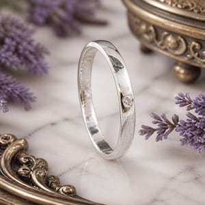 Puede incluir: Un anillo de plata pulida con un pequeño diamante. El anillo se encuentra sobre lavanda morada y una superficie de mármol, con un joyero de estilo antiguo en el fondo.