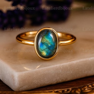 May include: A gold ring with an oval labradorite gemstone. The gemstone displays iridescent blue, green, and yellow hues. The ring is set on a white marble surface. The brand name "Latelierbijoux" is visible in the background.