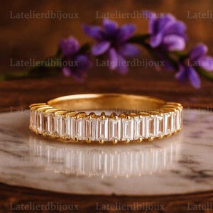 May include: A gold band ring with a row of rectangular, clear gemstones. The ring is set on a reflective surface, with purple flowers in the background. The ring is a piece of jewelry.