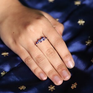 Puede incluir: Un anillo de plata con un grupo de piedras preciosas azules y moradas. El anillo se muestra en una mano sobre un fondo azul oscuro texturizado. Las piedras preciosas son de varios tama&ntilde;os y formas, creando un efecto brillante.
