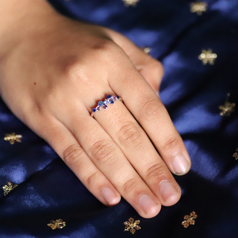 Puede incluir: Un anillo de plata con un grupo de piedras preciosas azules y moradas. El anillo se muestra en una mano sobre un fondo azul oscuro texturizado. Las piedras preciosas son de varios tama&ntilde;os y formas, creando un efecto brillante.
