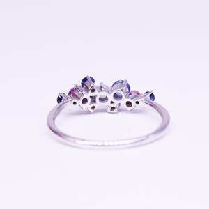 Puede incluir: Un anillo de plata con un grupo de piedras preciosas redondas en tonos azules y rosas. El anillo tiene una banda fina y pulida, y las piedras est&aacute;n engastadas juntas, creando un dise&ntilde;o floral o de estrella. El anillo est&aacute; sobre un fondo blanco.