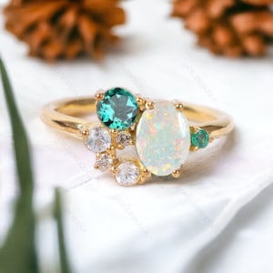 Könnte beinhalten: Ein goldener Ring mit einem großen ovalen Opal-Edelstein, zwei kleineren smaragdgrünen Edelsteinen und einem Cluster kleinerer klarer Edelsteine.