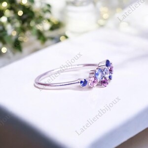 Puede incluir: Un anillo de plata con un grupo de peque&ntilde;as piedras preciosas de colores. El anillo presenta una banda delicada y est&aacute; adornado con piedras azules, moradas y rosas. Las piedras preciosas est&aacute;n dispuestas en un dise&ntilde;o floral o en racimo.
