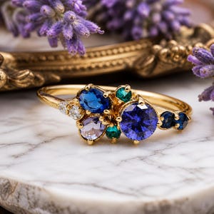 Puede incluir: Un anillo de oro con un grupo de piedras preciosas azules, verdes y moradas. El anillo tiene una banda delicada y está colocado sobre una superficie de mármol. Las piedras preciosas incluyen una gran piedra azul redonda y varias piedras más pequeñas.
