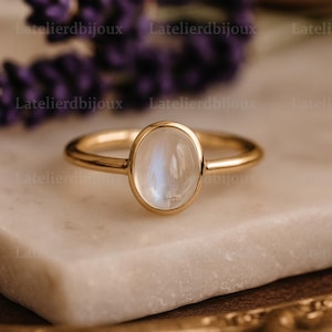 May include: A gold ring with an oval, iridescent moonstone. The ring is set on a white marble surface, with a blurred background of purple flowers and a gold-colored frame. The ring's band is thin and polished.