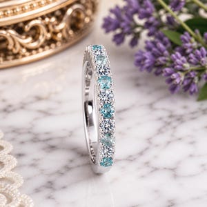 Op de afbeelding: Een zilveren ring met afwisselend ronde, lichtblauwe en heldere edelstenen. De ring is geplaatst op een marmeren oppervlak met een vage achtergrond met een gouden doos en paarse bloemen.