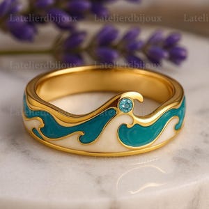 Könnte beinhalten: Ein goldener Ring mit Wellenmuster. Der Ring hat ein türkis-weißes Emaille-Wellenmuster um das Band, mit einem kleinen blauen Edelstein. Der Ring wird auf einer Marmoroberfläche präsentiert.