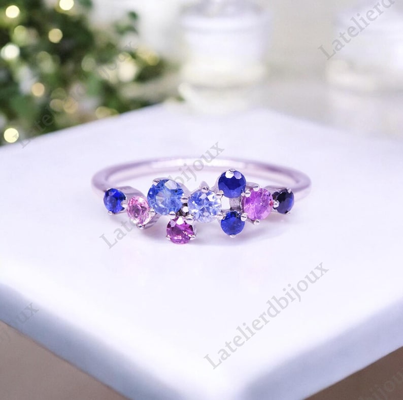 Puede incluir: Un anillo de plata con un grupo de piedras preciosas de colores. El anillo presenta una variedad de piedras redondas en tonos azules, morados y rosas. Las piedras est&aacute;n engastadas juntas, creando un dise&ntilde;o &uacute;nico y llamativo.