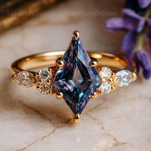 May include: A gold ring with a large, kite-cut gemstone displaying blue and purple colours. The ring is embellished with smaller, clear gemstones. The ring is set against a marble surface.