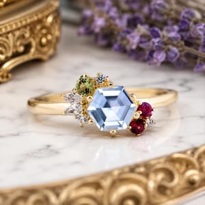 May include: A gold ring featuring a central hexagonal light blue gemstone, accented by a cluster of smaller stones including a round olive green gem, two round deep pink gems, and several clear white marquise and round cut accent stones.
