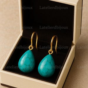 Puede incluir: Un par de pendientes turquesa en forma de l&aacute;grima con ganchos dorados, presentados en una caja de joyer&iacute;a beige con interior negro. Los pendientes tienen una superficie texturizada. El texto "Latelierdbijoux" es visible.